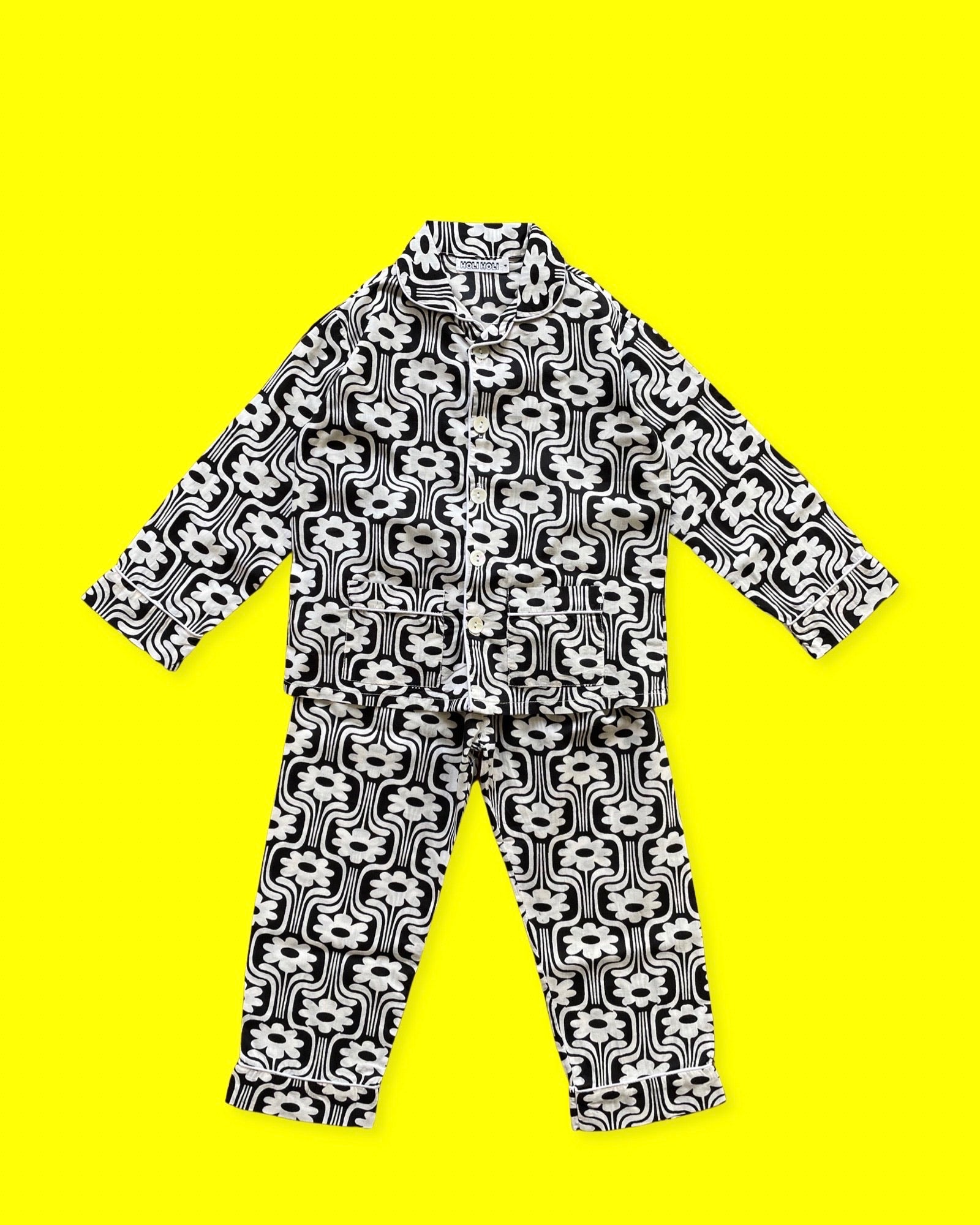 Ce pyjama enfant est un ensemble composé d'un haut boutonné à manches longues et d'un pantalon à taille élastique. L'imprimé est rétro à fleurs blanches sur fond noir, et un liseré blanc. La photo le présente sur un fond jaune.