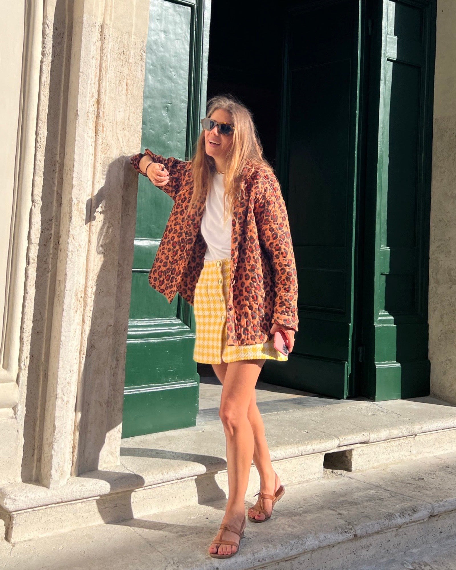 Le mannequin porte la chemise de chambre à imprimé léopard par-dessus un tee-shirt blanc et une jupe jaune et blanche à carreaux. Le mannequin est sur des marches au soleil.