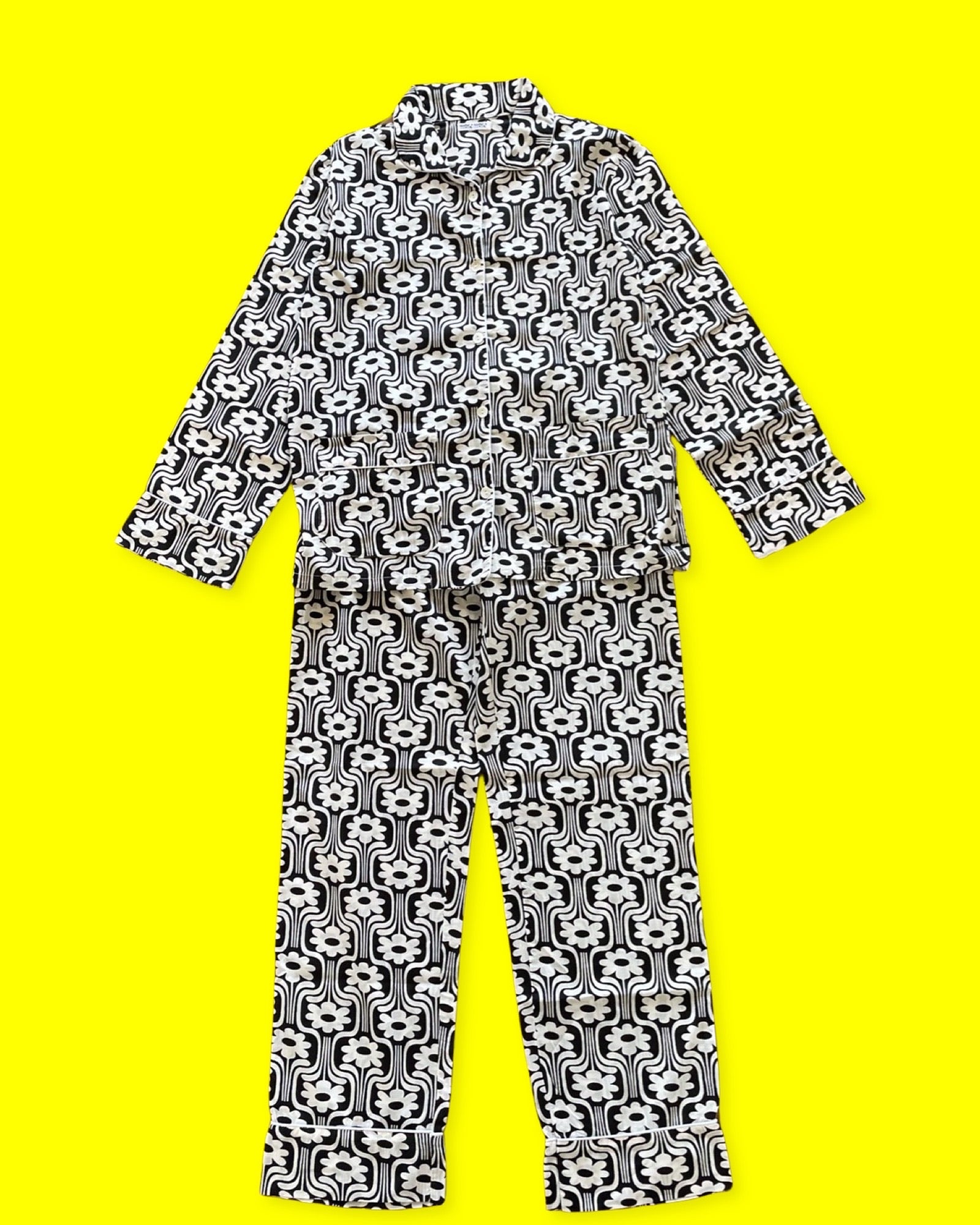 Notre pyjama pour femme 70's est un ensemble composé d'un haut boutonné à manches longues avec deux poches et d'un pantalon à taille élastique et à lien. L'imprimé est rétro vec des fleurs blanches sur fond noir, et le liseré est blanc. La photo le présente sur fond jaune.