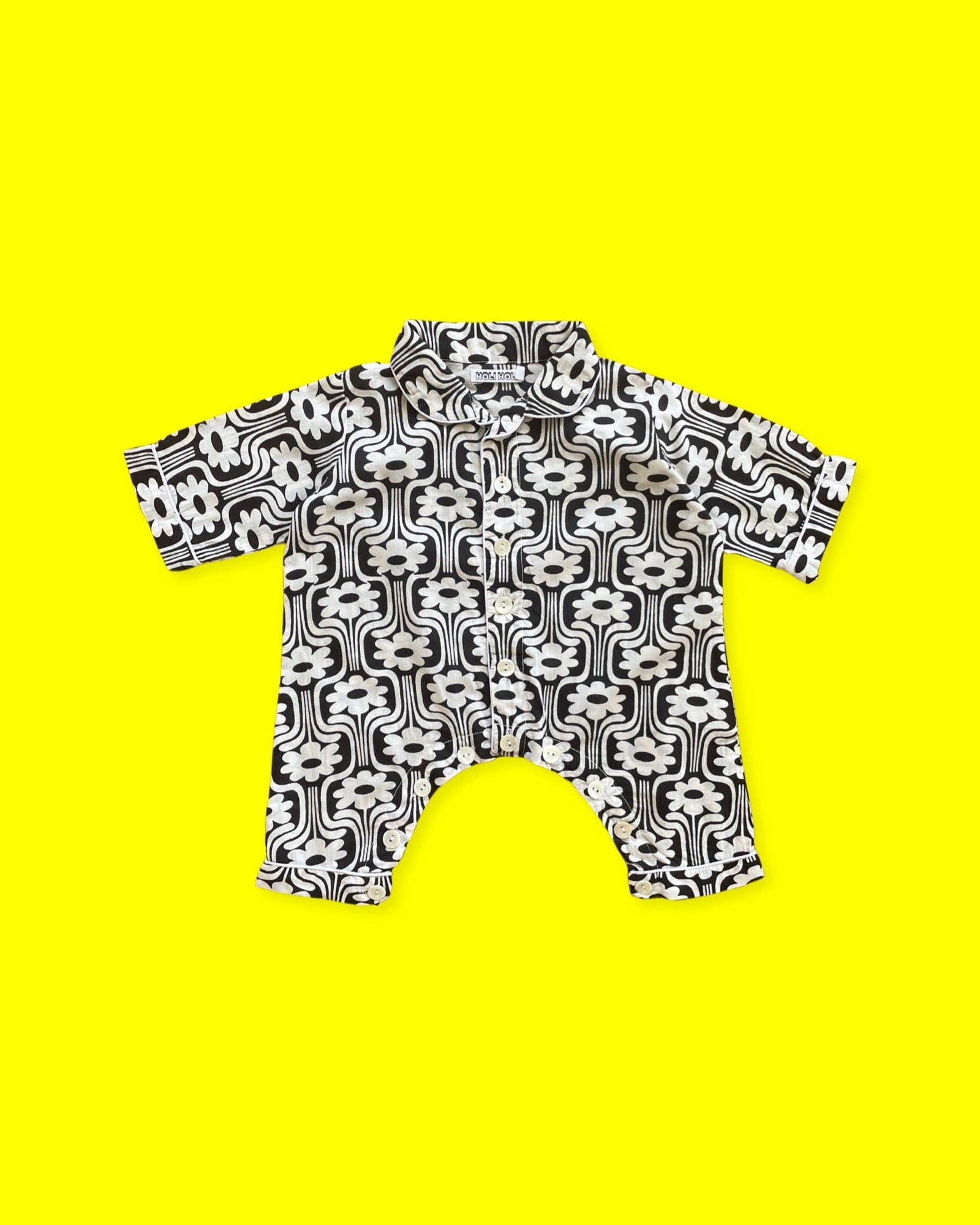 Ce pyjama pour bébé est une grenouillère à manches longues fermée à l'avant et à l'entrejambe par des petits boutons en nacre. Il a un imprimé rétro à fleurs blanches sur fond noir, et un liseré blanc, et un liseré blanc. Le pyjama est présenté sur un fond jaune.