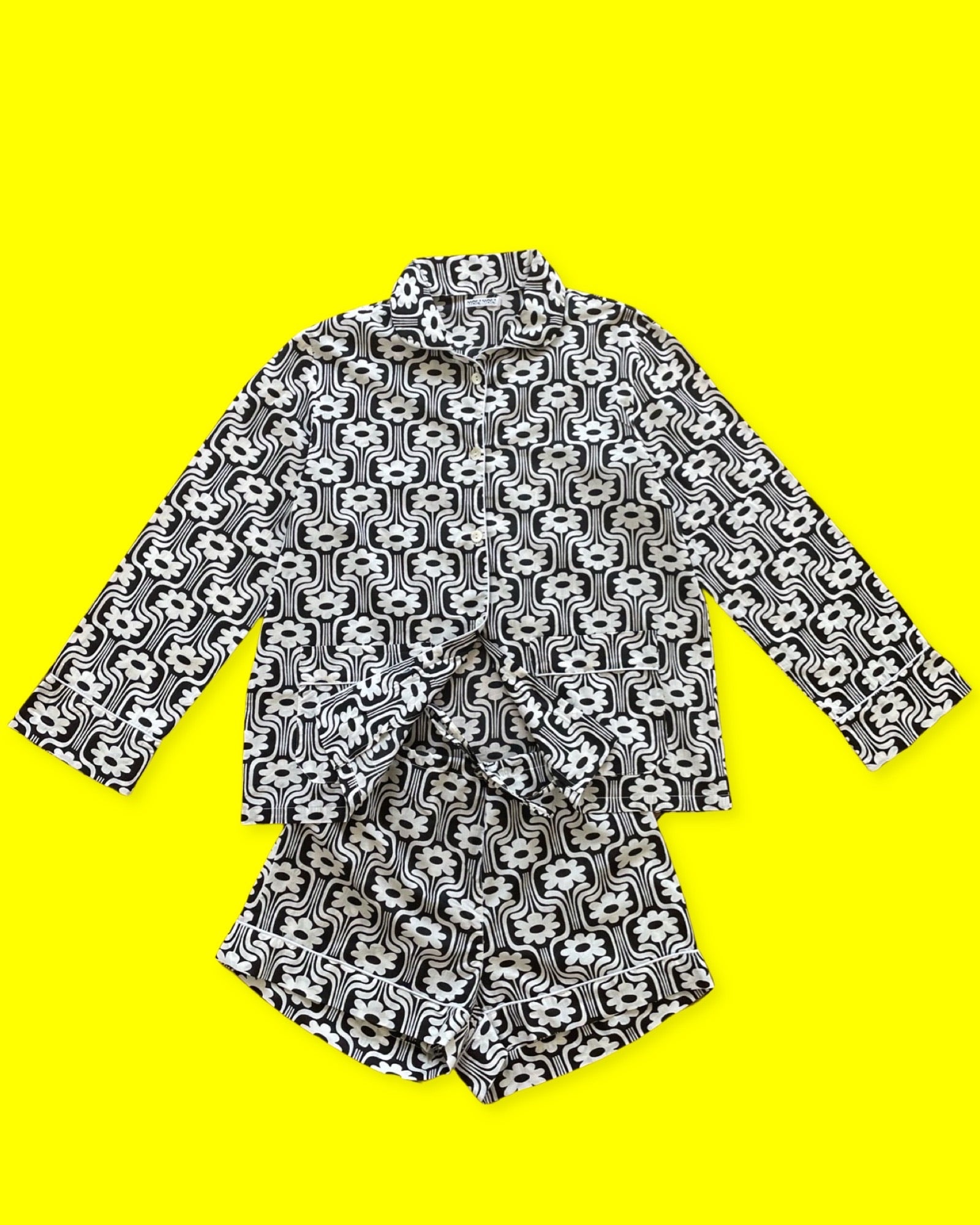 Ce pyjama court pour femme est un ensemble composé d'un haut boutonné à manches longues avec deux poches sur les côtés en bas de la veste et d'un short à taille élastique et à lien. L'imprimé est rétro à fleurs blanches sur fond noir, et un liseré blanc. La photo le présente sur un fond jaune.