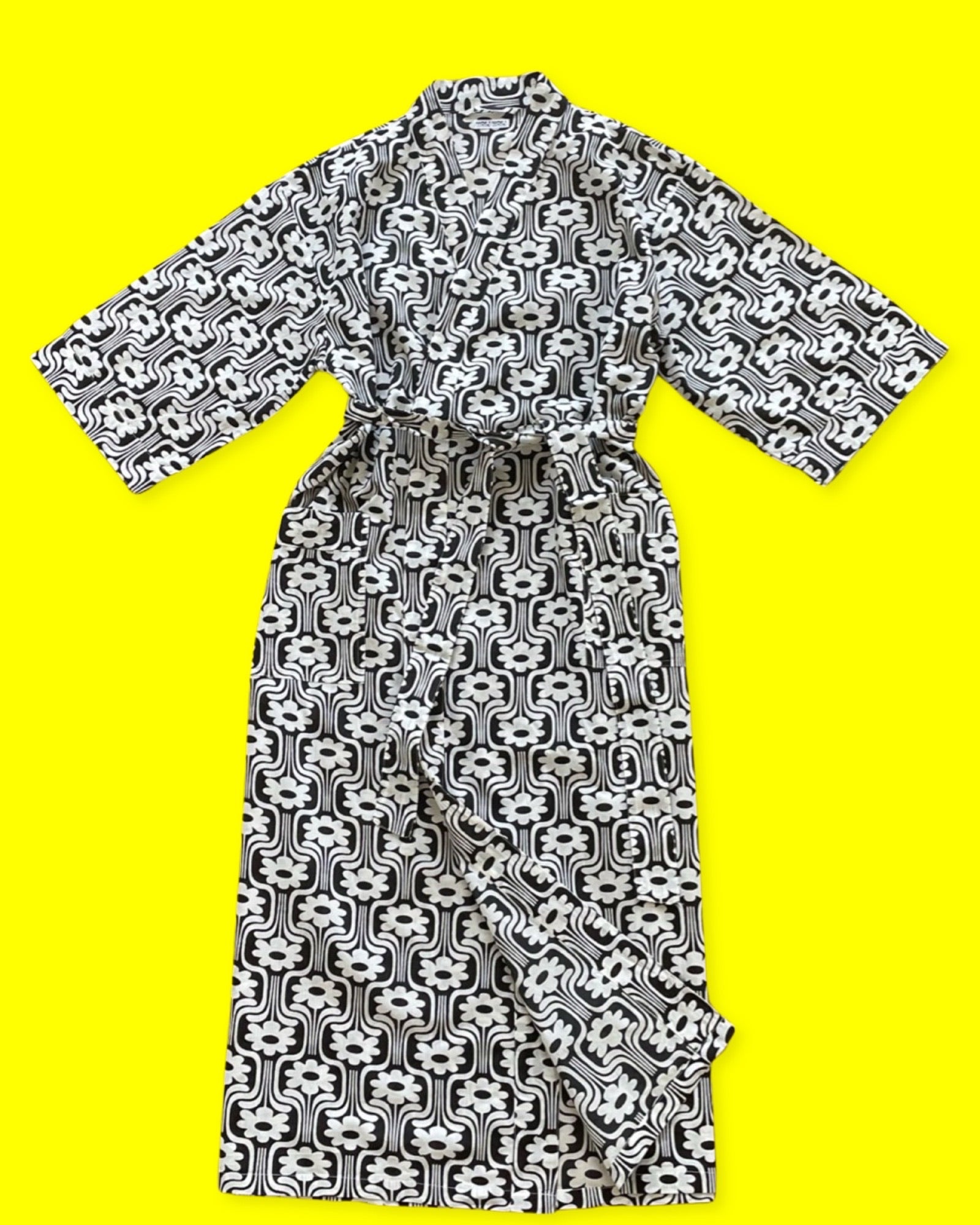 Notre sortie de lit est un kimono long à manches mi-longues, avec deux poches et fermée par une ceinture, le tout en coton fin à imprimé rétro à fleurs blanches sur fond noir. La photo la présente sur un fond jaune. 